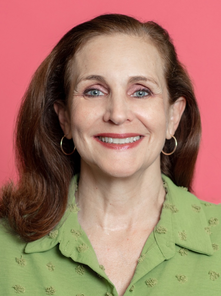 A woman with light skin, wearing a green top and hoop earrings, is smiling against a pink background. She has long, straight brown hair.